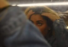 A teen girl is running her fingers through her hair while looking at her reflection in a slightly blurry mirror. Her arm covers one of her eyes and the bottom of her face, leaving only the top of her face visible.