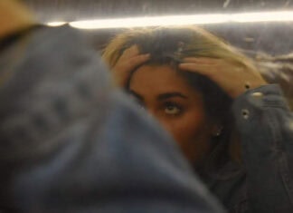 A teen girl is running her fingers through her hair while looking at her reflection in a slightly blurry mirror. Her arm covers one of her eyes and the bottom of her face, leaving only the top of her face visible.